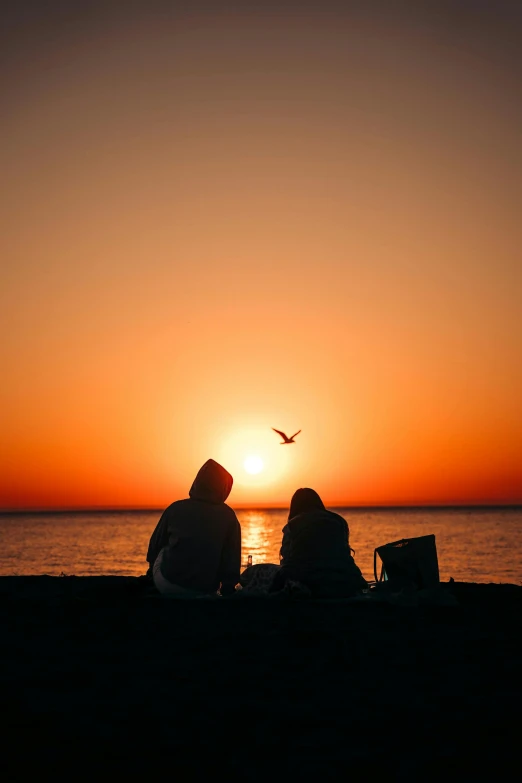 two people sit on the sand in front of a sunset