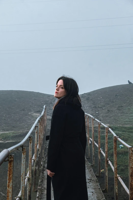 a woman in a long black coat stands alone on the bridge