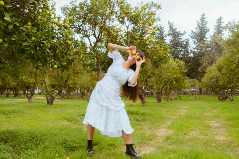 a woman in white dress holding up apples with trees behind her