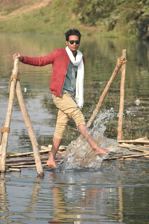 a man crosses a stream holding up a bunch of sticks and wearing a sweater