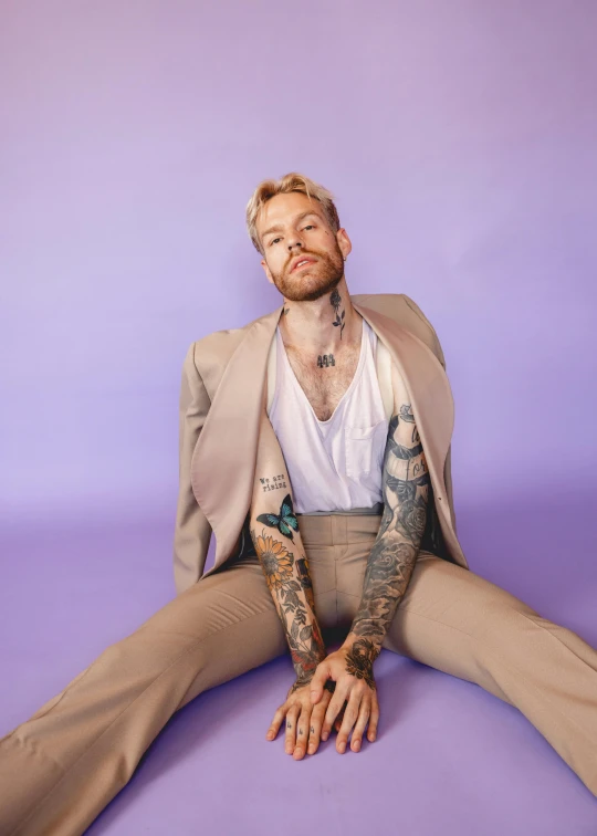 the tattooed man sits down in a suit and holds his arms out wide