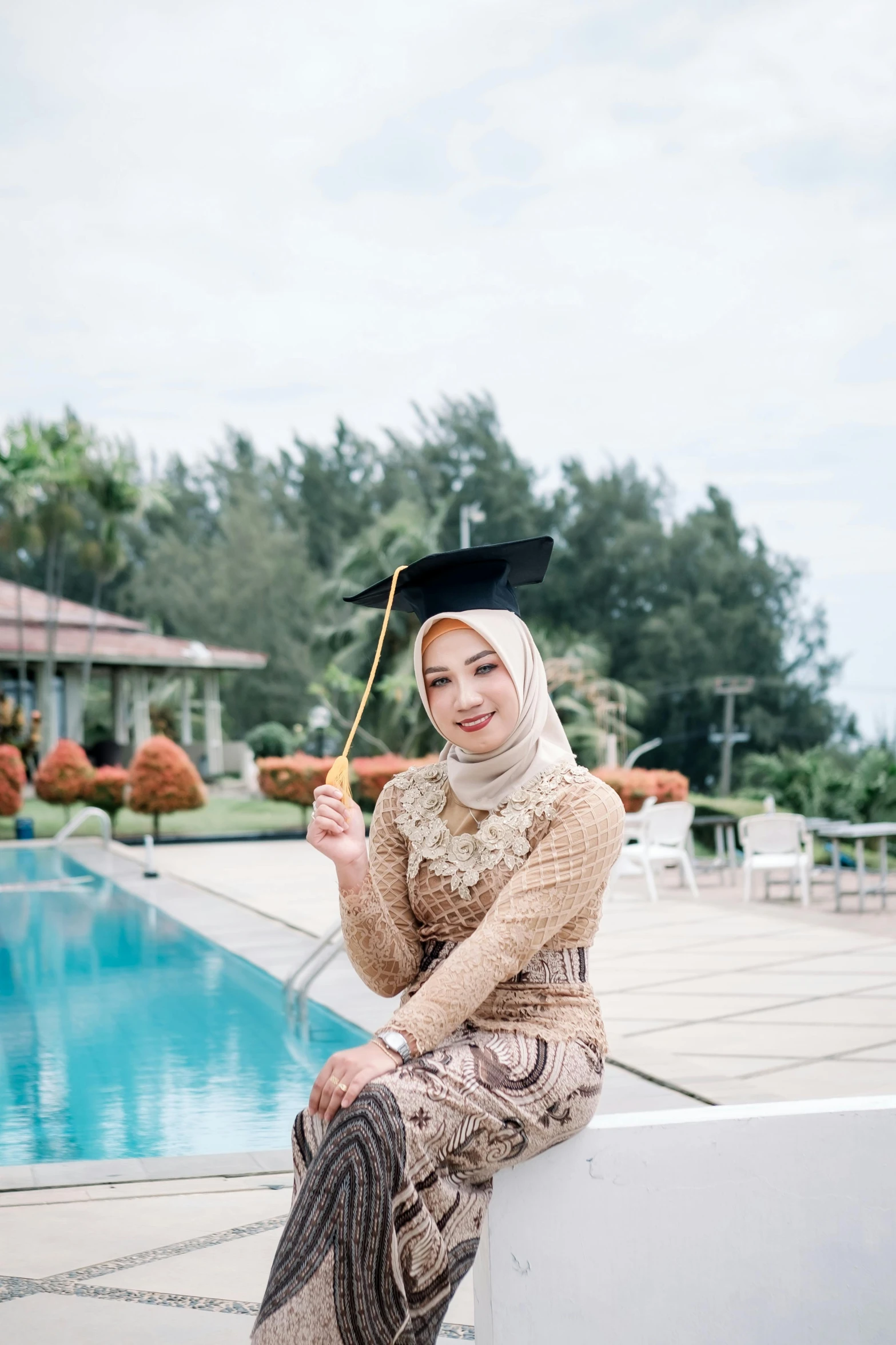 the young woman in a scarf and graduation gown is sitting by the pool