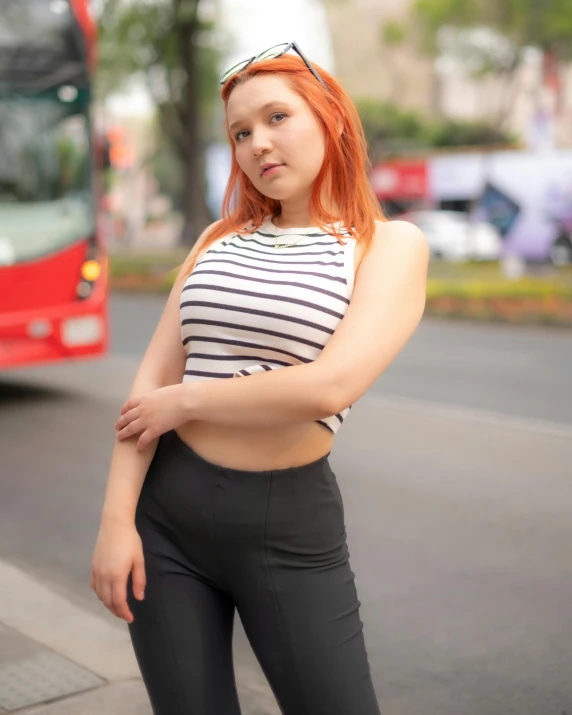 a girl wearing tight pants, a white and black striped top, and sunglasses stands near a red bus on a road