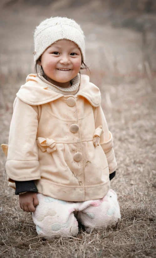 a little girl in a coat and hat sits in a field