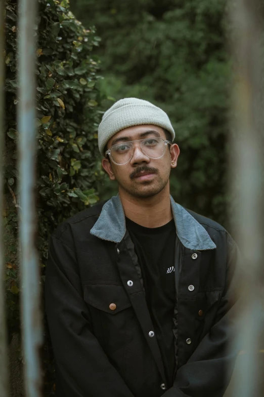 a man in a black jacket and cap stands against a fence