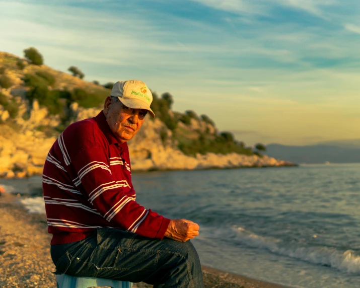 an old man wearing a baseball cap sitting on a skateboard at the beach
