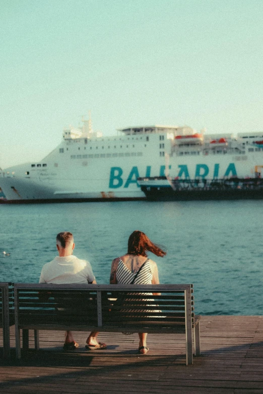 three people are sitting on a bench in front of the water and a large ship