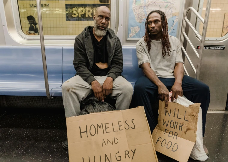 two homeless people sit on a subway station with sign'homeless and hungry '