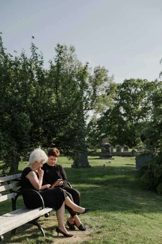 two women sit on a wooden bench in the middle of a grassy field