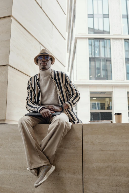 a man with a hat, striped jacket and white pants sits on the edge of a building