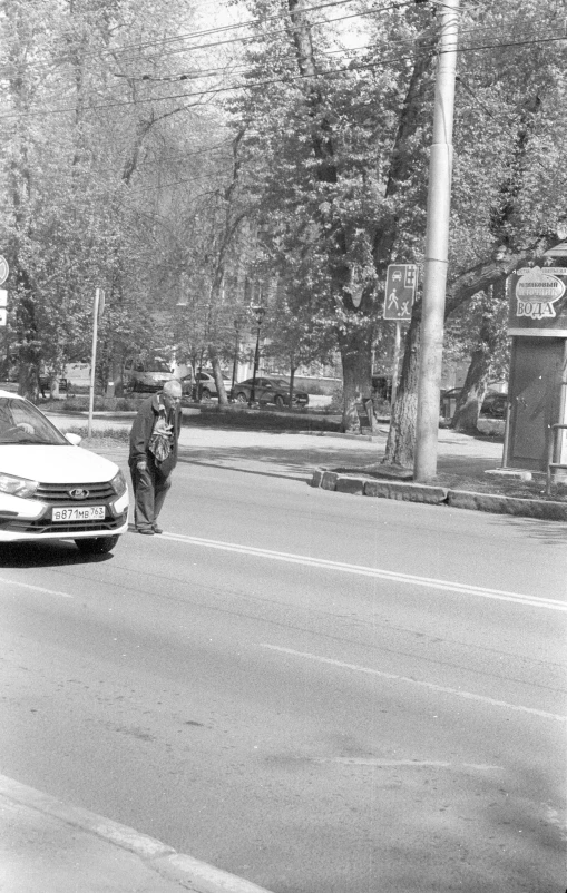 black and white image of a man on the side of a road with a parked car