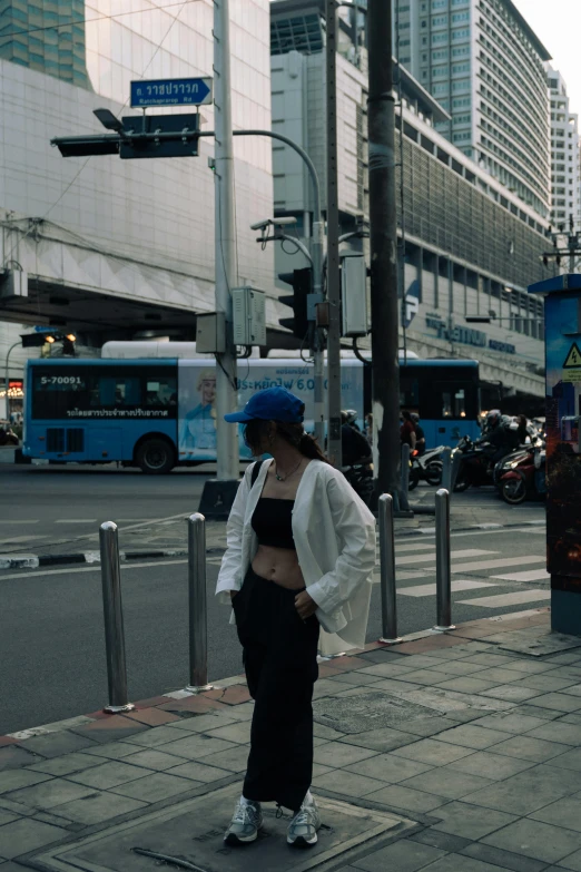 a woman in crop tops, jeans and white jacket with a blue visor stands at a crosswalk