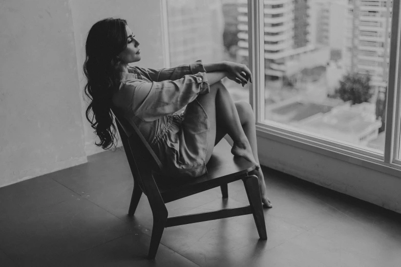 a woman in an oversized denim jacket is sitting on a chair looking out of a window