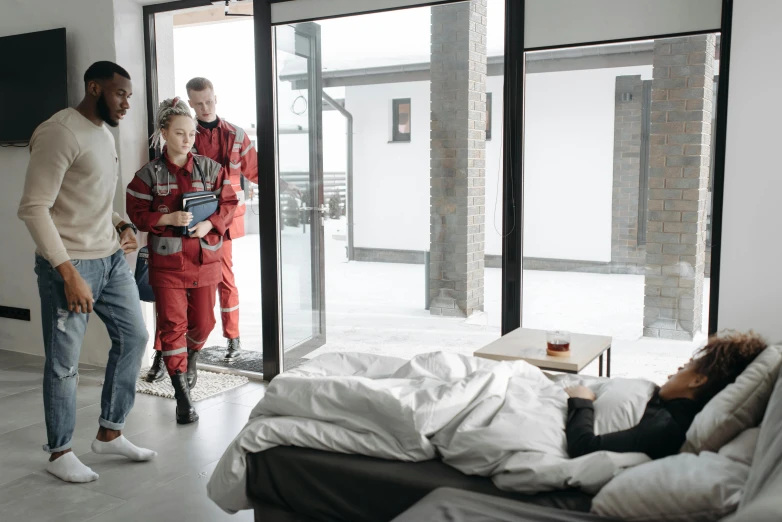 two men and a woman are standing near a bedroom window while an uninspected woman lays in bed behind them