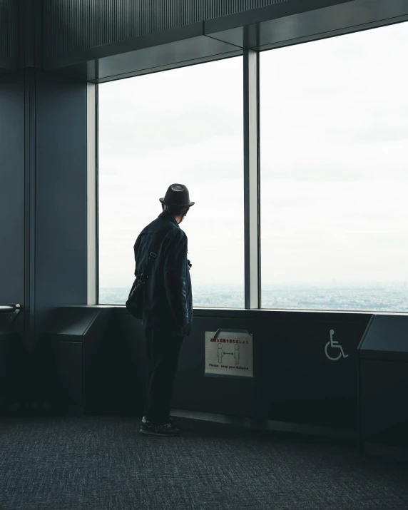 a man in a black suit and hat stands with his back towards a window