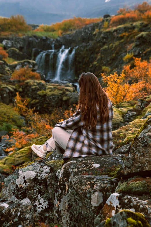 a woman with long red hair sitting on the rocks looking at a waterfall