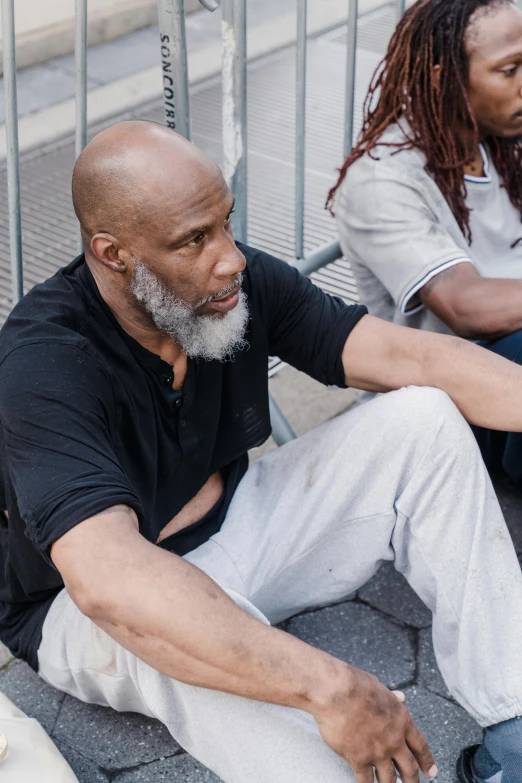 two men sitting and standing on top of a sidewalk