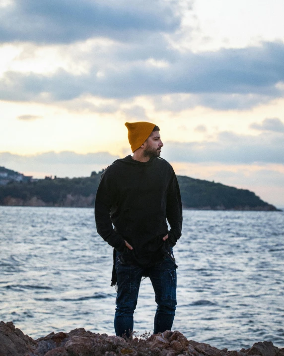 man with yellow beanie and jeans standing on rocky shoreline