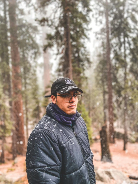 the man in black jacket and hat is posing by trees