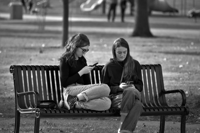 two women sitting on a park bench checking their phones