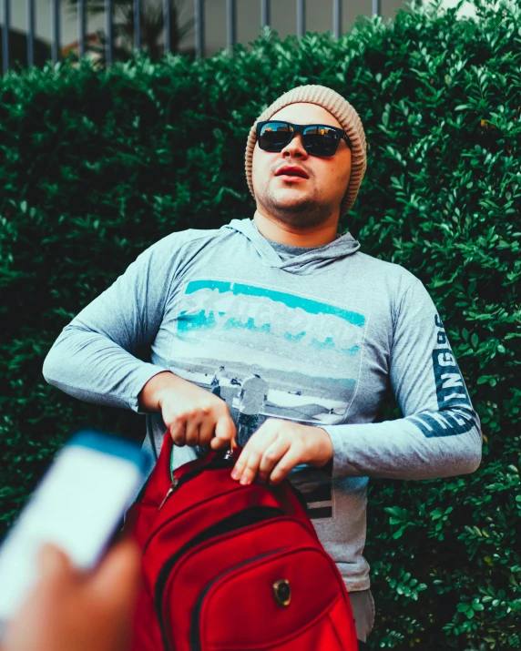 man in grey t shirt pulling red suitcase in front of bushes