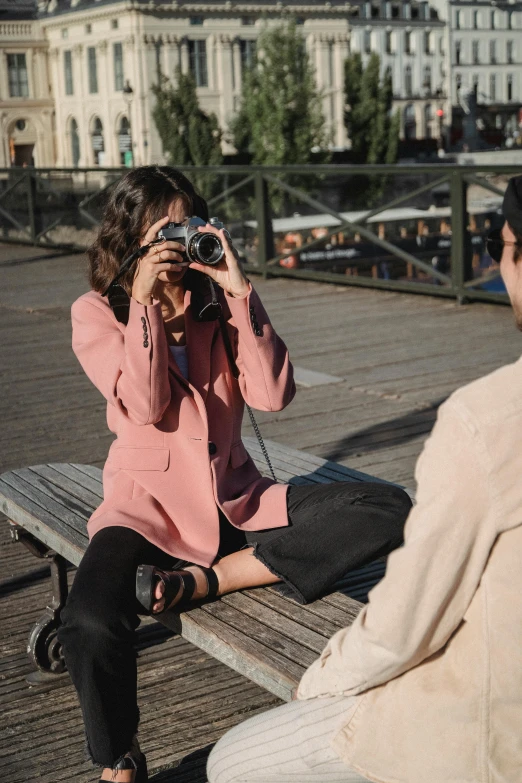 two young women sit on a bench near a building while one woman takes pos