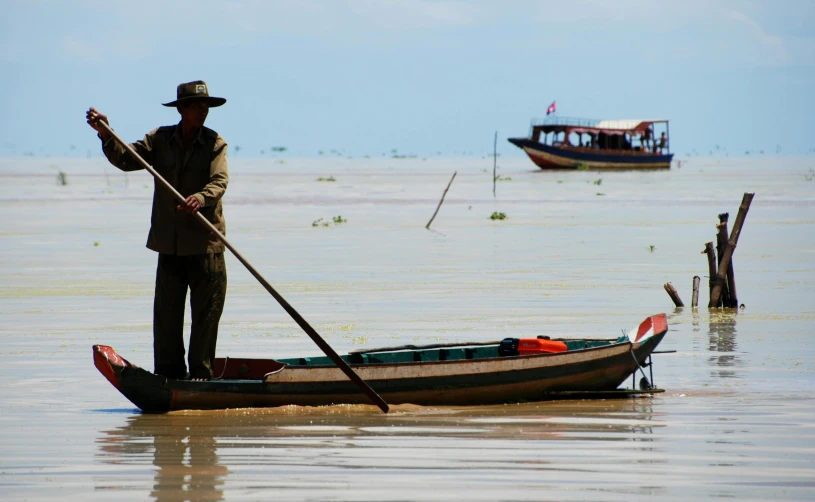 a man wearing a hat and long black cane stands on a small boat in shallow water