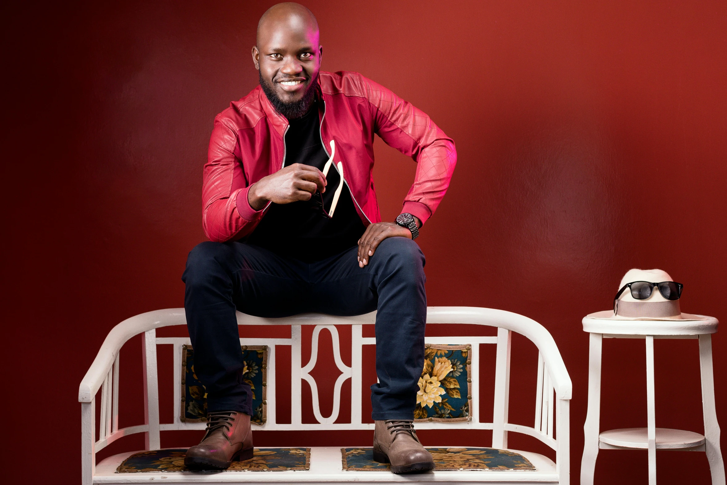 a man poses in front of a red wall with two small tables