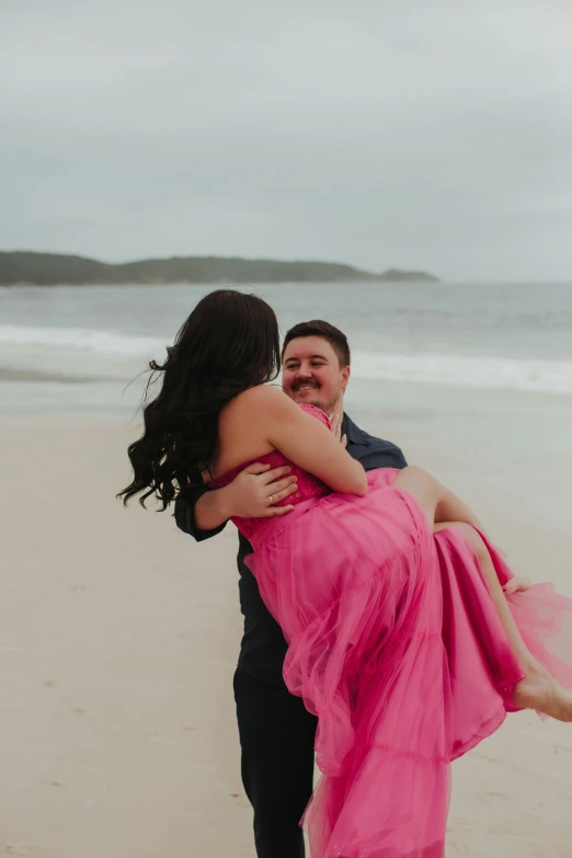 bride in pink dress carries groom on beach