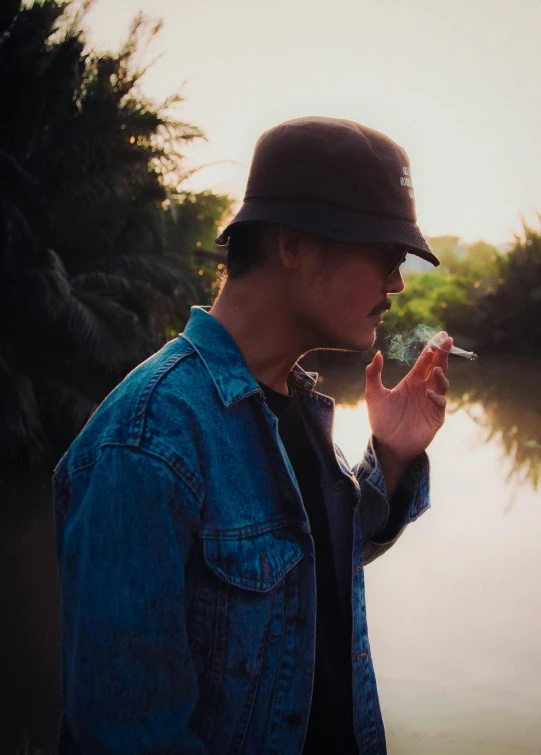 a man in a hat smoking a cigarette next to the river