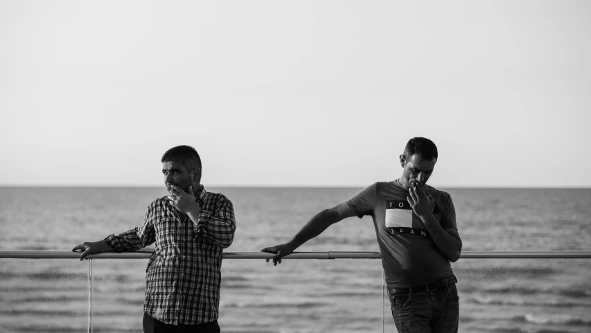 two young men stand near water and one talks on a cell phone