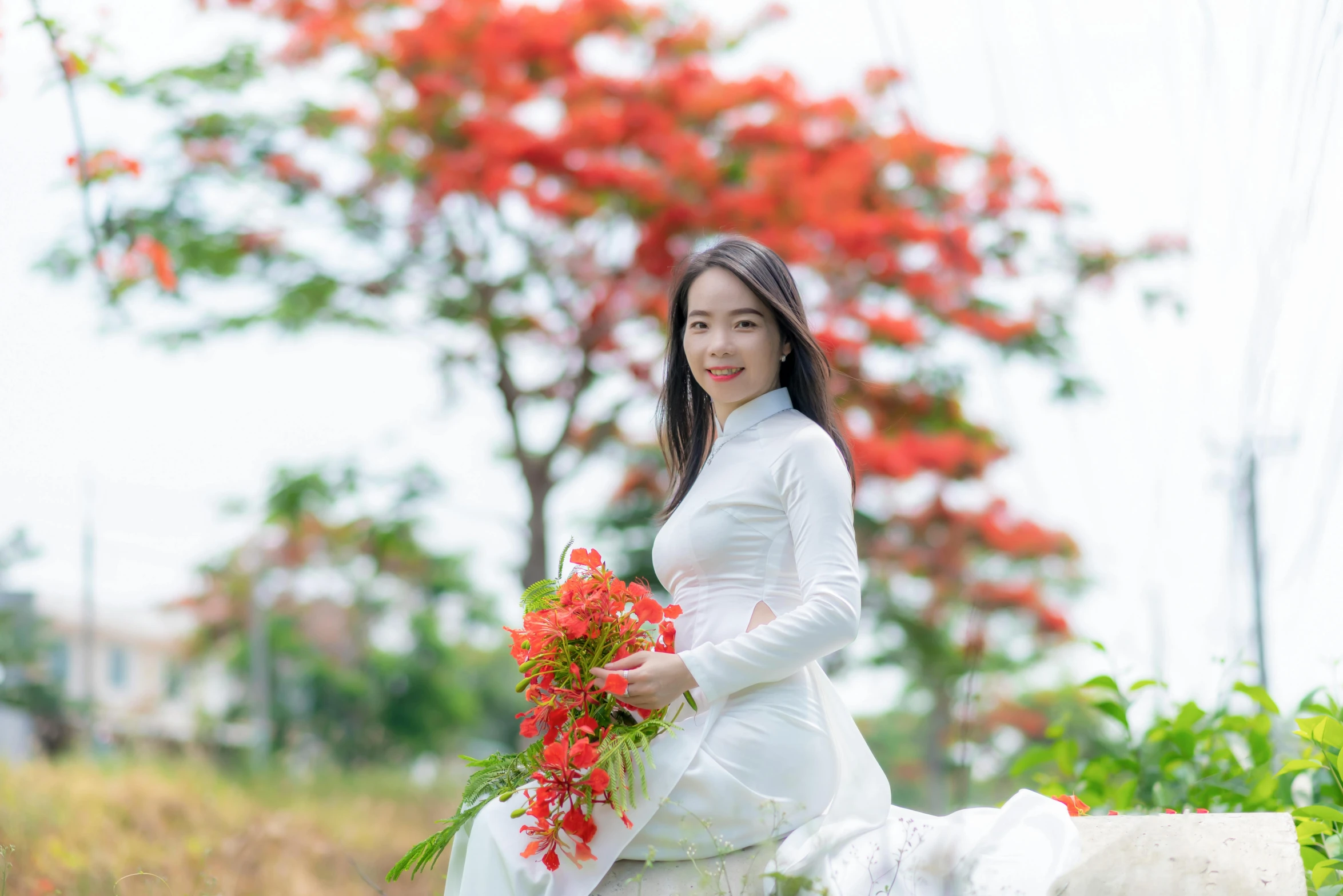 an asian woman dressed in a white long - sleeved gown holds a red flower bouquet and poses for the camera