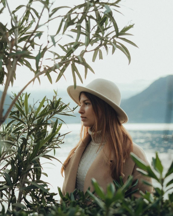 a girl in white hat standing near trees and water