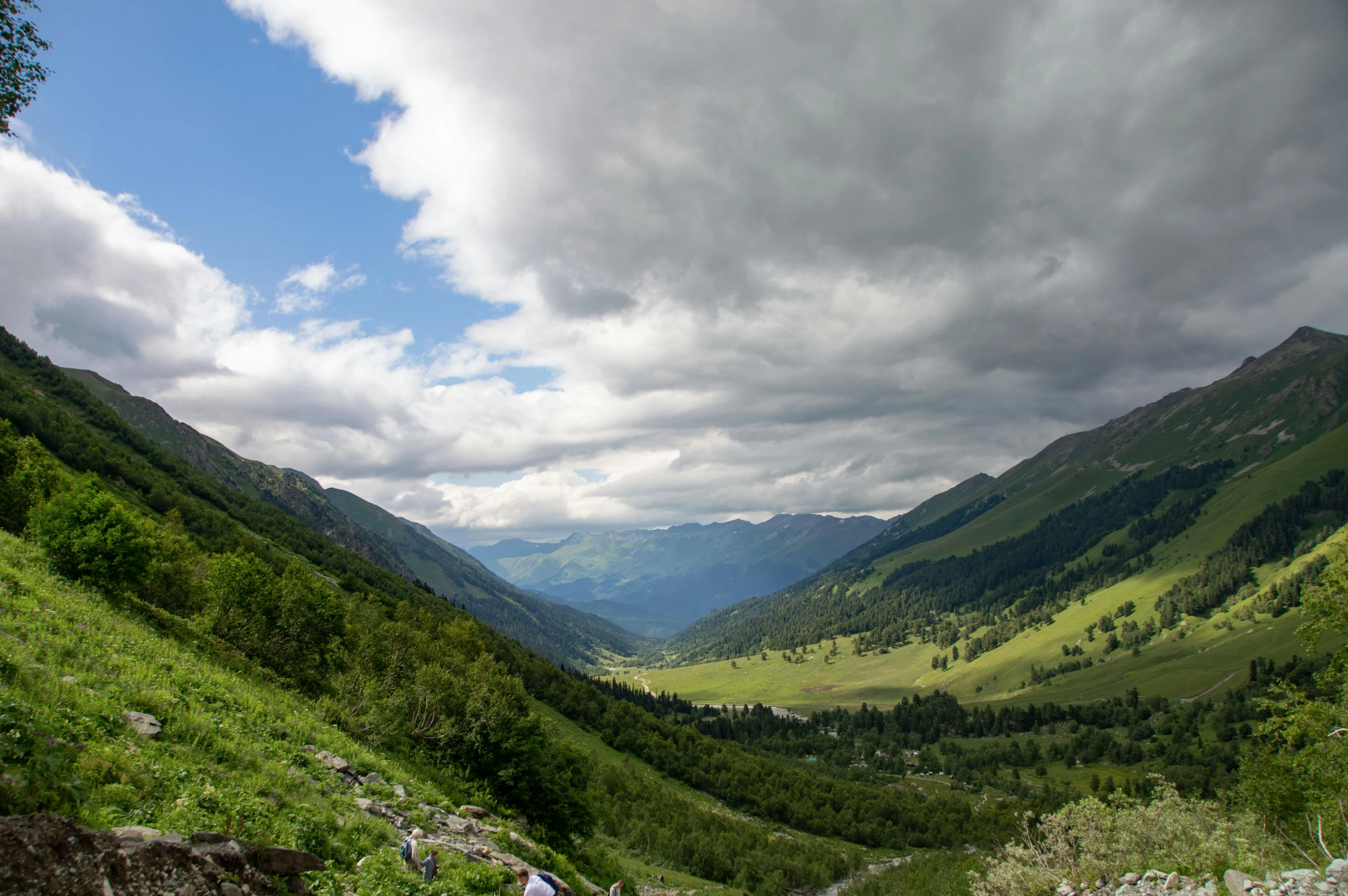 two people hiking up a mountain side, among green mountains