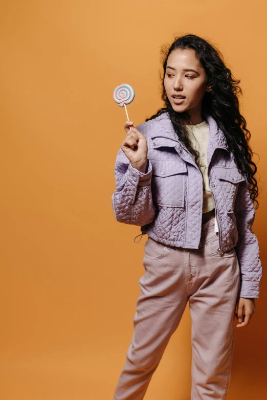 a girl in an unoned coat with an orange background holds a candy lollipop in front of her face