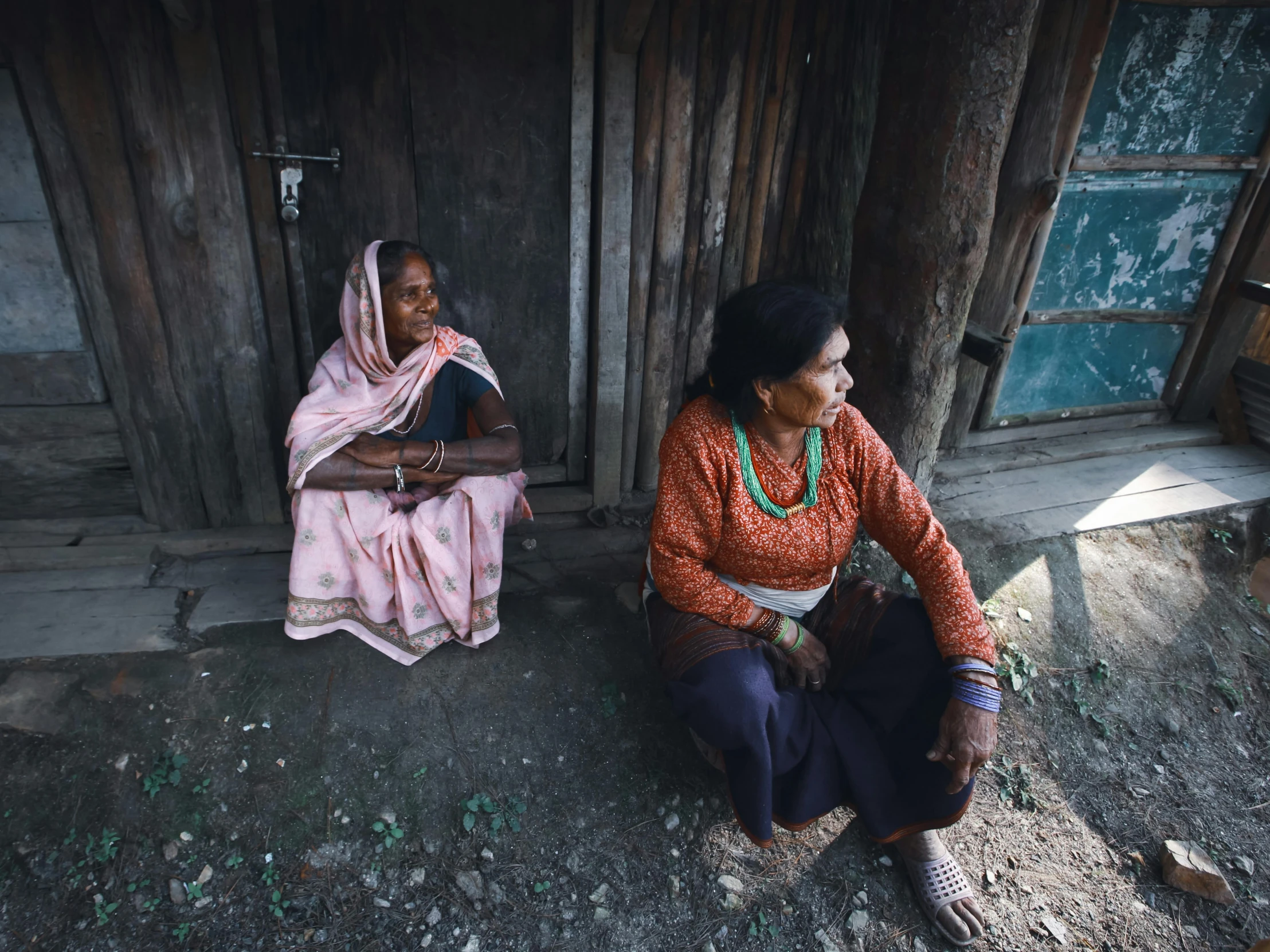 two women sit on the ground outside of their wooden houses