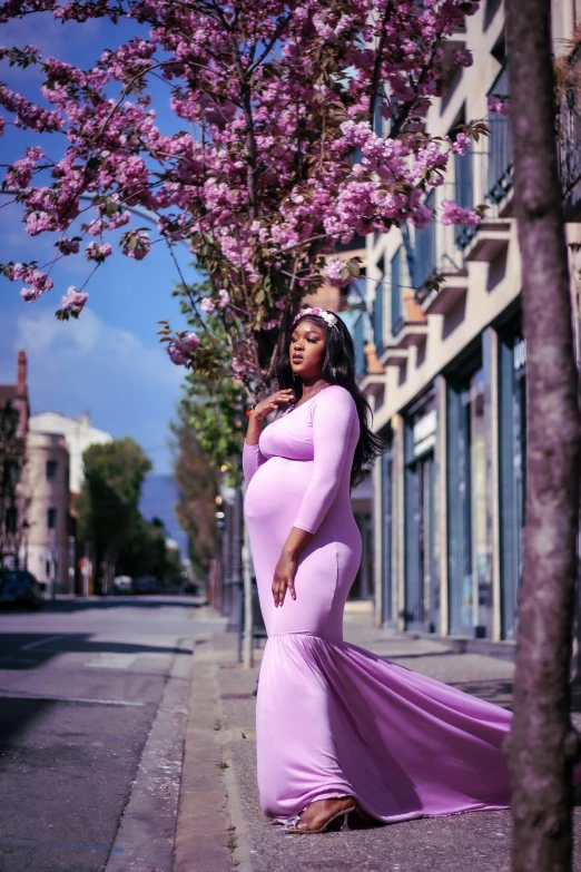 a pregnant lady is leaning on the tree with her hand in her pocket and standing on a sidewalk