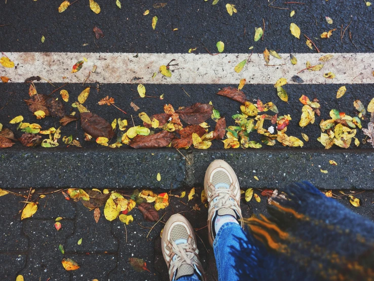a person in blue jeans and white sneakers standing near an intersection with leaves on the pavement