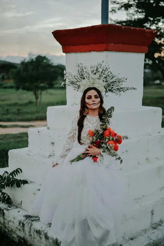 a woman wearing a dress and holding a bouquet sitting on a stone pillar with roses in front of her