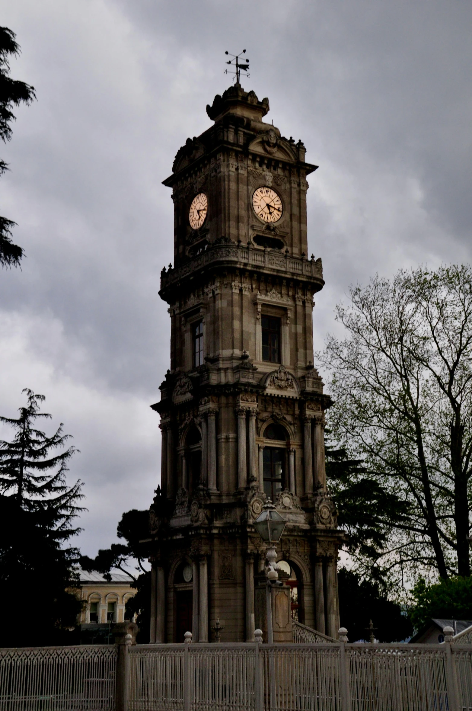 a stone clock tower with a large bell is shown in front of a fence and trees