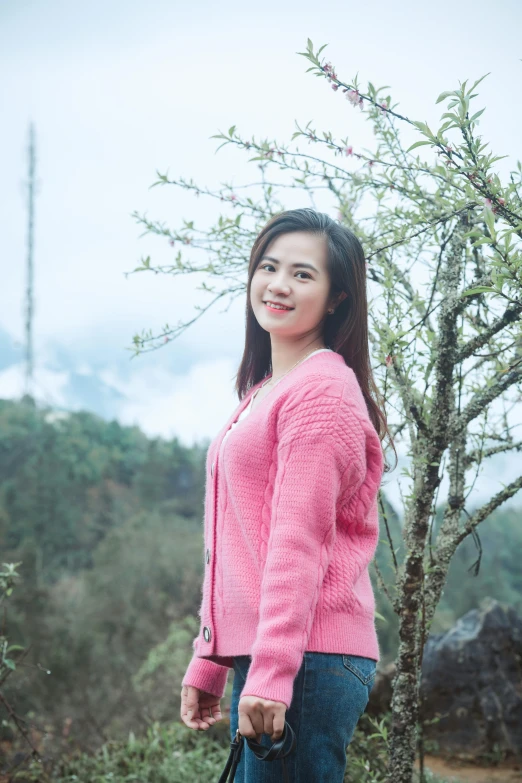a woman wearing a pink sweater and blue jeans poses in front of a small tree