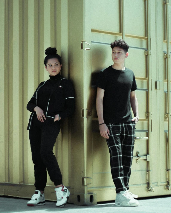 two people in black outfits standing against a wall