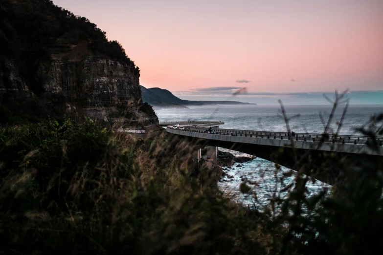 a view from the shoreline at sunset, with a bridge over water and people on it