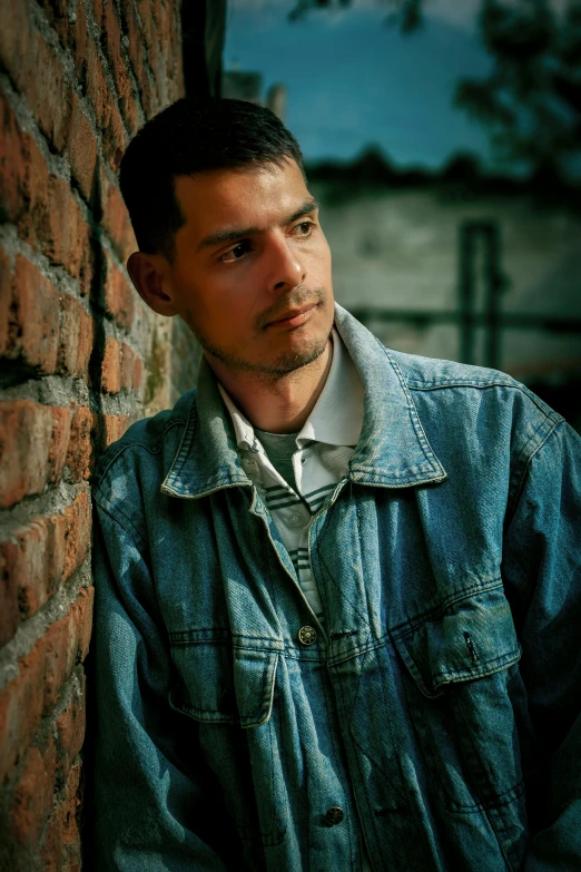 a guy leans against the wall wearing a blue denim jacket