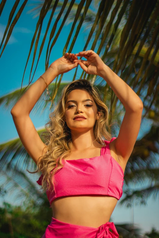 woman in pink cropped top holding hand over head, under palm tree