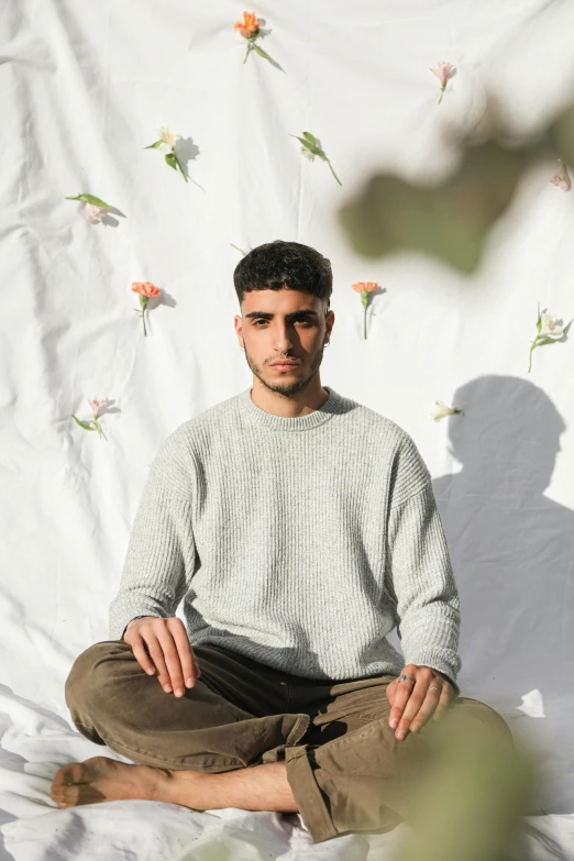 a man sitting on a white sheet with flowers on the wall behind him