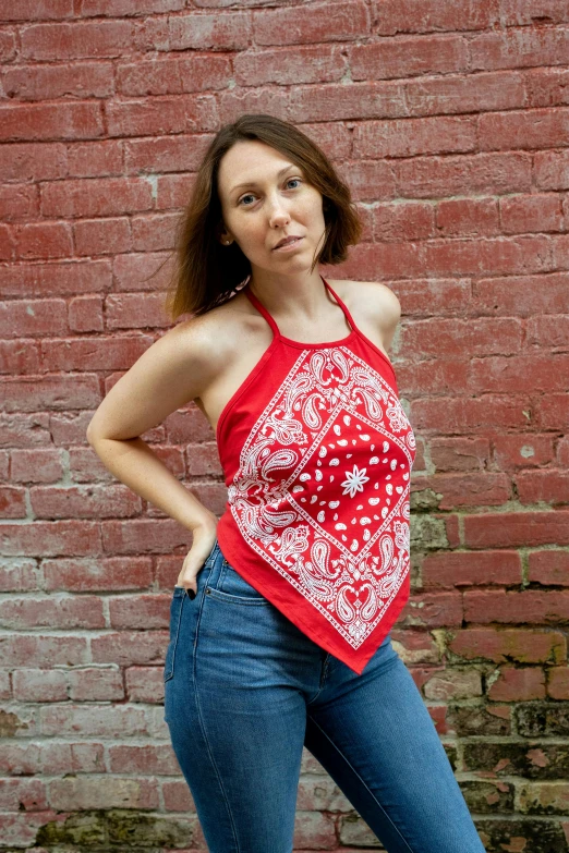 a woman standing against a brick wall wearing blue jeans and a red top with an interesting paisley print