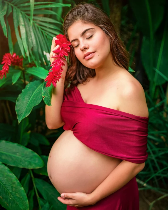 a pregnant girl in a red dress with her hand on her cheek, in front of green foliage