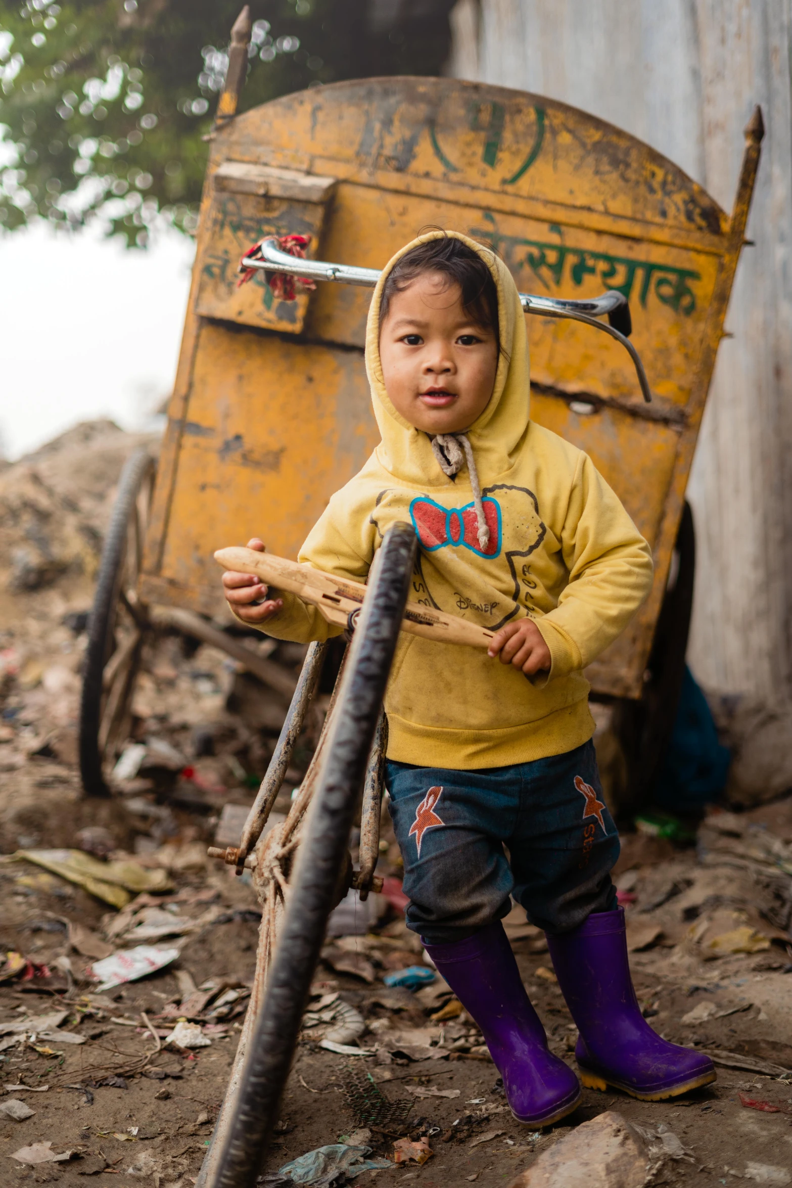 a small child wearing a yellow sweatshirt and rubber boots stands next to a pile of trash in a yard with an orange wheelbarrow