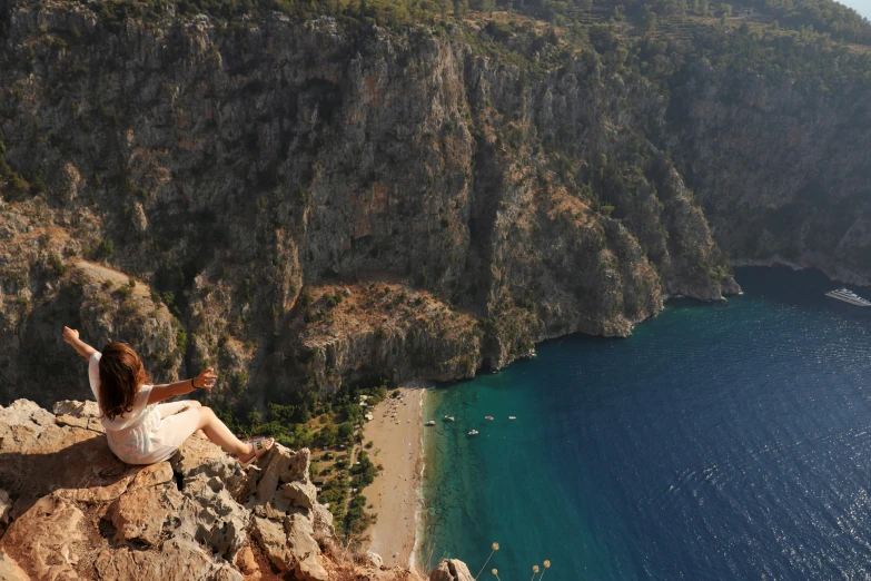 a woman sitting on the edge of a cliff looking over a beach